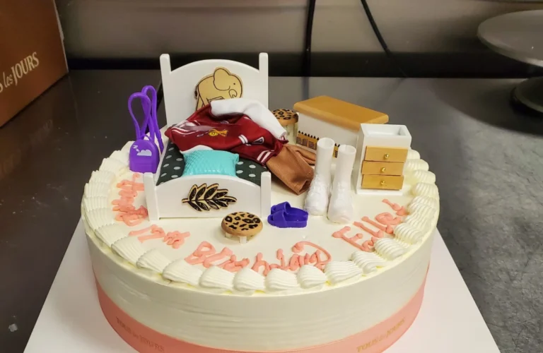 Custom birthday cake decorated with a miniature bed, books, and study-themed items on top, with the message “Happy Birthday” written in icing. Freshly made custom cake from TOUS les JOURS Iowa in Des Moines.